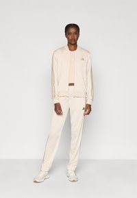 Σετ αθλητικών ρούχων σε ανοιχτό μπεζ χρώμα με ζακέτα μπότερ με φερμουάρ και ταιριαστό παντελόνι. Διαθέτει διακριτικές χρυσές λεπτομέρειες και πλαϊνές ρίγες. Περιλαμβάνει ροζ μπλούζα από μέσα.