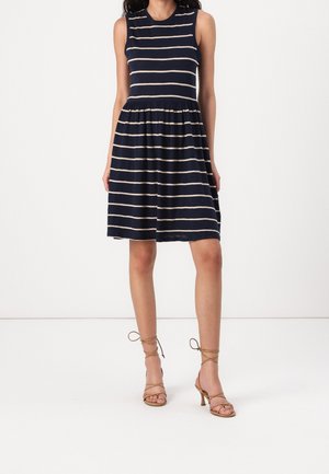 Femme portant une robe bleu marine sans manches avec des rayures horizontales blanches et des sandales à talons hauts à lanières couleur sable, debout devant un fond blanc.