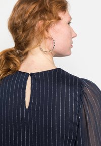 Blusa azul-marinho com riscas verticais prateadas, tecido texturizado, mangas ligeiramente bufantes e uma pequena abertura em forma de buraco de chave nas costas, com fecho de botão.