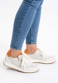 Weiße perforierte Sneakers mit orangen Heel-Akzenten. Besitzt eine Schnürung vorne und eine strukturierte Sohle. Kombiniert mit blauen Skinny Jeans.