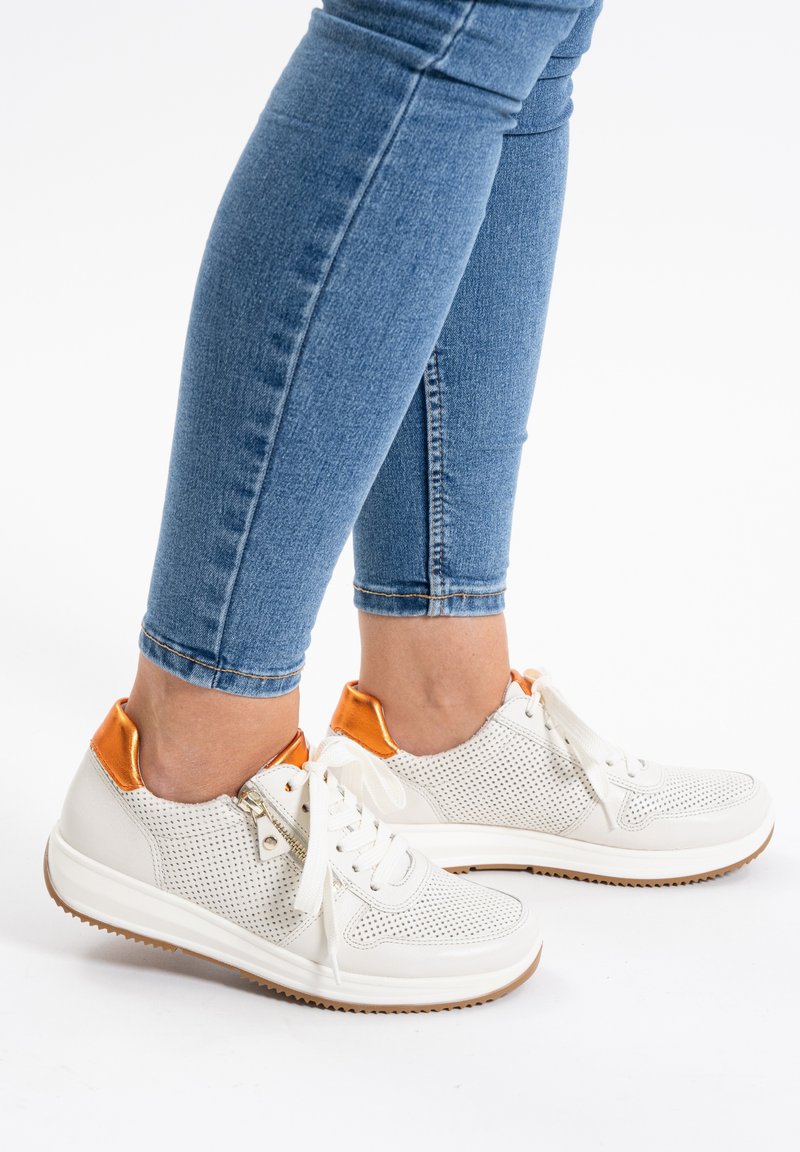 Weiße perforierte Sneakers mit orangen Heel-Akzenten. Besitzt eine Schnürung vorne und eine strukturierte Sohle. Kombiniert mit blauen Skinny Jeans.