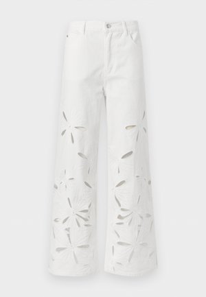 Žena nosiaca biely tielkový top bez rukávov, biele široké nohavice s vyrezávanými kvetinovými vzormi, hnedé baleríny a okrúhle slnečné okuliare, stojaca s rukami v vreckách.