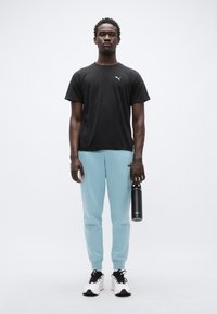 Μαύρο αθλητικό μπλουζάκι, ανοιχτό μπλε φόρμα, και μαύρα αθλητικά παπούτσια· κρατώντας μαύρο παγούρι νερού· απλό φόντο, στάση όρθια.