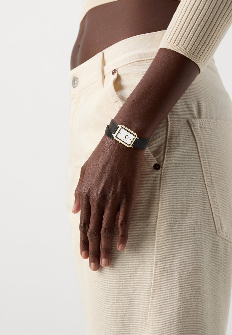 Montre en cuir noir avec un cadran blanc rectangulaire, accents dorés, portée au poignet, associée à un pantalon en velours côtelé crème.