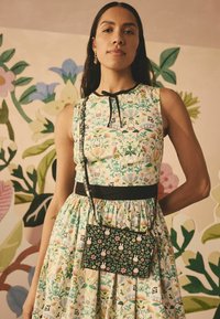 Floral-patterned sleeveless dress with a fitted waist and black belt; paired with a small black bag adorned with colorful floral designs.