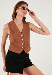 Vestă maro din piele întoarsă, cu decolteu în V, șase nasturi din lemn și două buzunare frontale. Asortată cu o fustă neagră, modelul poartă ochelari de soare rotunzi.