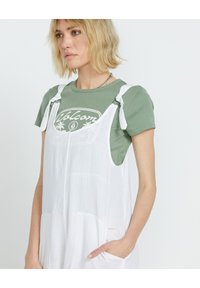 Abito lungo in lino bianco con tasche laterali, spalline regolabili, indossato sopra una maglietta verde con logo stampato. Tessuto morbido.