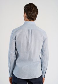Uomo di spalle che indossa una camicia a maniche lunghe azzurro chiaro con bottoni e pantaloni scuri, su uno sfondo bianco semplice.