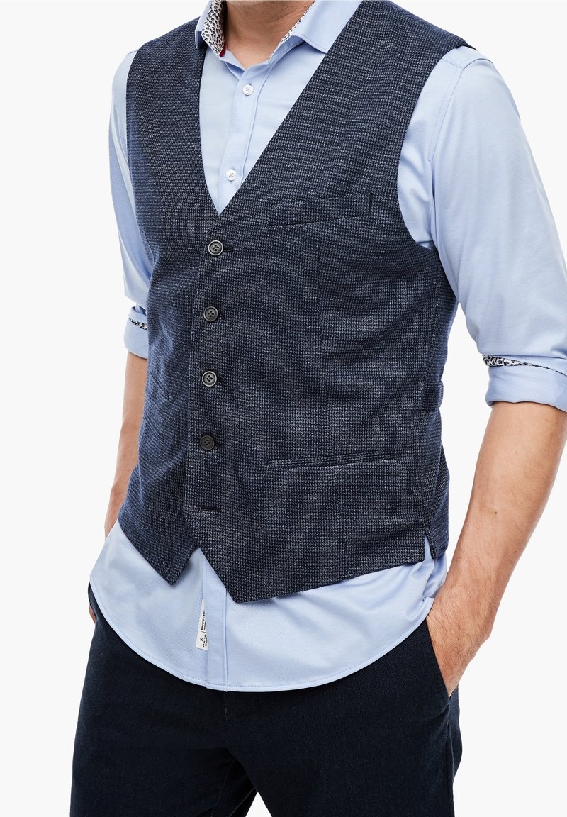Dunkelblaues gemustertes Weste mit V-Ausschnitt, fünf Knöpfen, einer einzelnen Brusttasche, getragen über einem hellblauen Hemd mit hochgekrempelten Ärmeln.