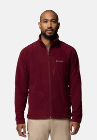 Giacca in pile bordeaux con colletto alto, chiusura con zip e due tasche laterali. Logo Columbia ricamato sul petto. Texture morbida.