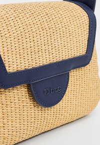 Borsa in vimini color sabbia con pattina e tracolla in ecopelle blu navy, dotata di una chiusura rotonda con il marchio "O bag". Superficie intrecciata testurizzata.
