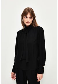 Blusa nera realizzata in tessuto fluido, con maniche lunghe, un fiocco al collo e dettagli a bottone sui polsini. Tessuto morbido, design elegante.