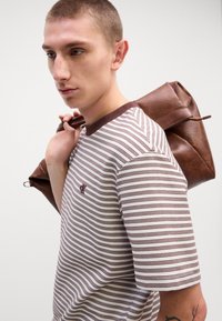 Brązowa koszula w paski z krótkim rękawem i brązowym kołnierzykiem, z małym logo na klatce piersiowej. Brązowa skórzana torba przewieszona przez ramię.