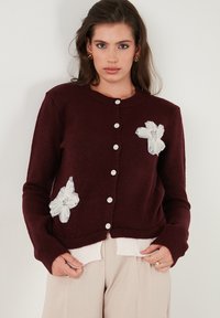 Burgundský pletený cardigan s bielymi kvetinovými aplikáciami, okrúhlym výstrihom, prednými gombíkmi a rebrovanými manžetami, preložený cez svetlý top.