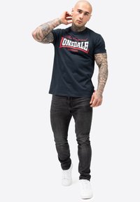 Marineblaues T-Shirt mit weißem und rotem "Lonsdale London"-Druck, Rundhalsausschnitt, kurze Ärmel; kombiniert mit schwarzen Jeans und weißen Turnschuhen.