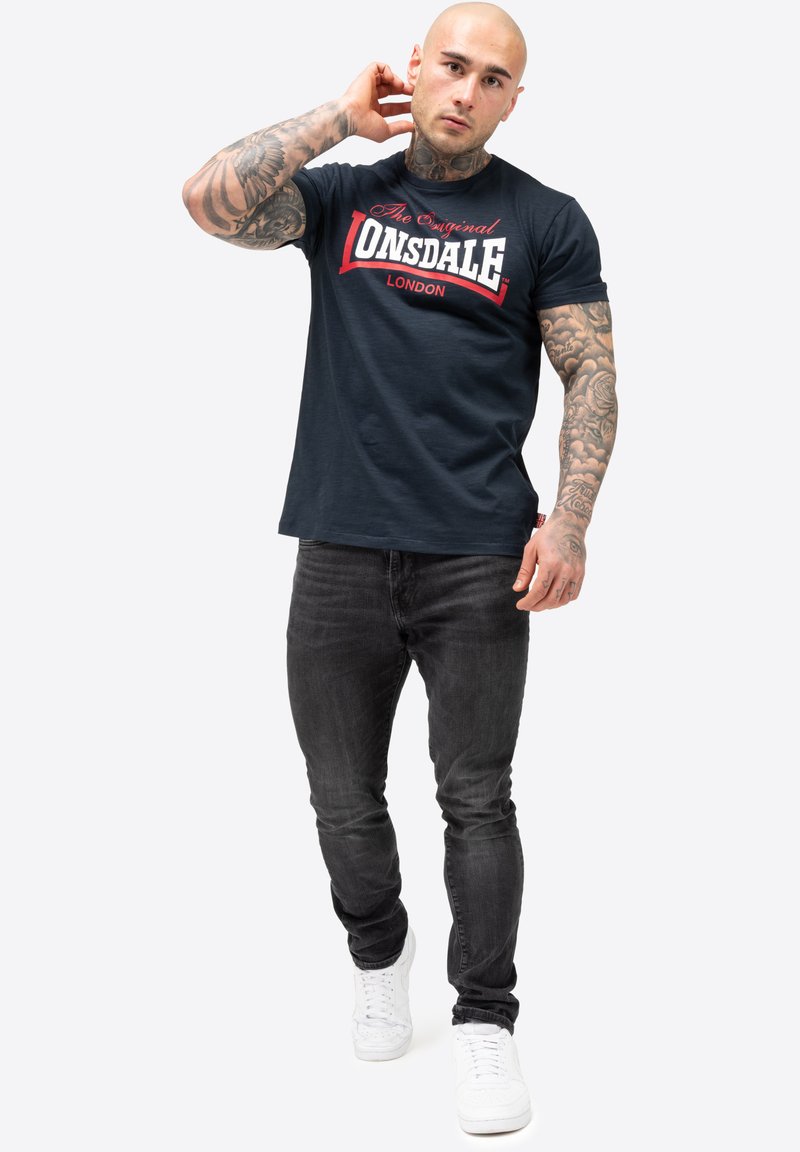 Marineblaues T-Shirt mit weißem und rotem "Lonsdale London"-Druck, Rundhalsausschnitt, kurze Ärmel; kombiniert mit schwarzen Jeans und weißen Turnschuhen.