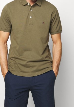 Man draagt een olijfgroen poloshirt met korte mouwen en een klein logo op de borst, gecombineerd met marineblauwe broek, handen in de zakken, op een witte achtergrond.