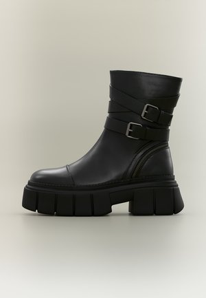 Bottine en cuir noir avec semelle épaisse, deux sangles ajustables avec boucles métalliques et détail d'une fermeture éclair latérale.