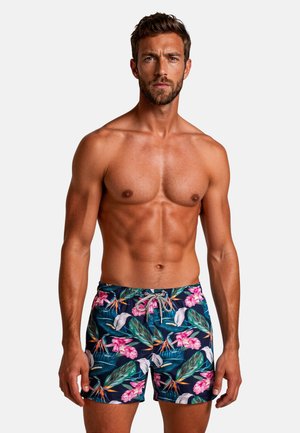 Muskelöser Mann mit kurzen braunen Haaren, der bunte, florale Badeshorts trägt, steht vor einem schlichten Hintergrund und schaut mit ernstem Gesichtsausdruck nach vorne.