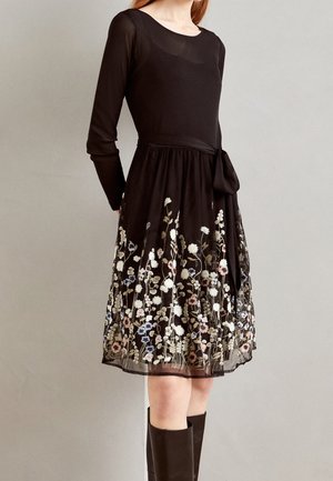 Femme portant une robe noire à manches longues avec une jupe brodée de fleurs et une ceinture à la taille, associée à des bottes noires montantes au genou.