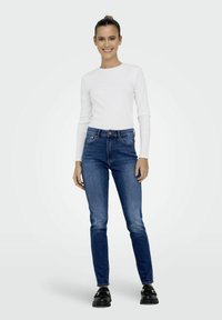 Balta ilgomis rankovėmis palaidinė, aukštu liemeniu mėlyni džinsai, juodi batai; siauras džinsinis modelis su klasikinėmis kišenėmis ir siuvinėjimo detalėmis.