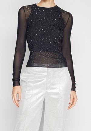 Czarna przejrzysta bluzka z długim rękawem ozdobiona drobnymi kryształkami, zestawiona z wysokim stanem błyszczących białych spodni, noszona przez stojącą osobę.