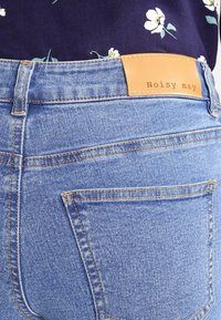Jeans de denim de color azul claro con una cintura alta, que presentan costuras naranjas y una etiqueta de cuero marrón claro con la inscripción "Noisy may" en la parte trasera.
