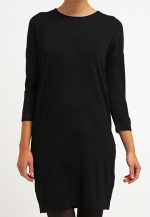 Kobieta ubrana w prostą, czarną sukienkę z długim rękawem sięgającą kolan, z okrągłym dekoltem, na białym tle.