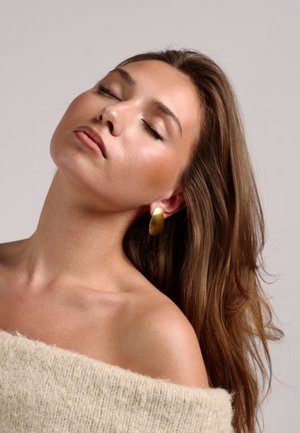 Gouden oorringen, gladde afwerking, ovale vorm, gedragen door een model met lang haar, gehuld in een zachte, lichte trui.