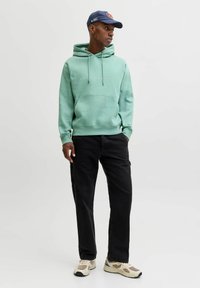 Svijetlozeleni dukserica s vezicama i prednjim džepom, uparen s crnim hlačama. Obuća ima krem i sivo dizajn. Kapa s logotipom je pričvršćena.