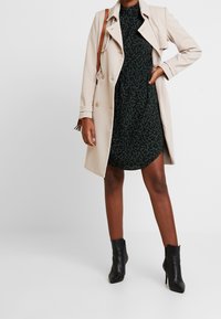 Beige trenchcoat över en mörkgrön klänning med svart blommönster. Svarta ankelskor, brun axelväska synlig. Släta tygtexturer.