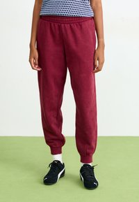 Bordeaux sweatpants med elastisk talje og manchetter, lavet af blødt stof. Sorte træningssko med hvide detaljer, stående på grønne gulve.