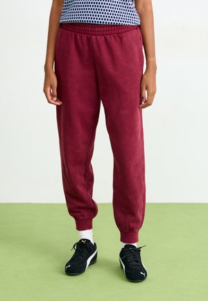 Burgunderfargede joggebukser med elastisk liv og mansjetter, laget av myk stoff. Svarte treningssko med hvite detaljer, stående på grønt gulv.