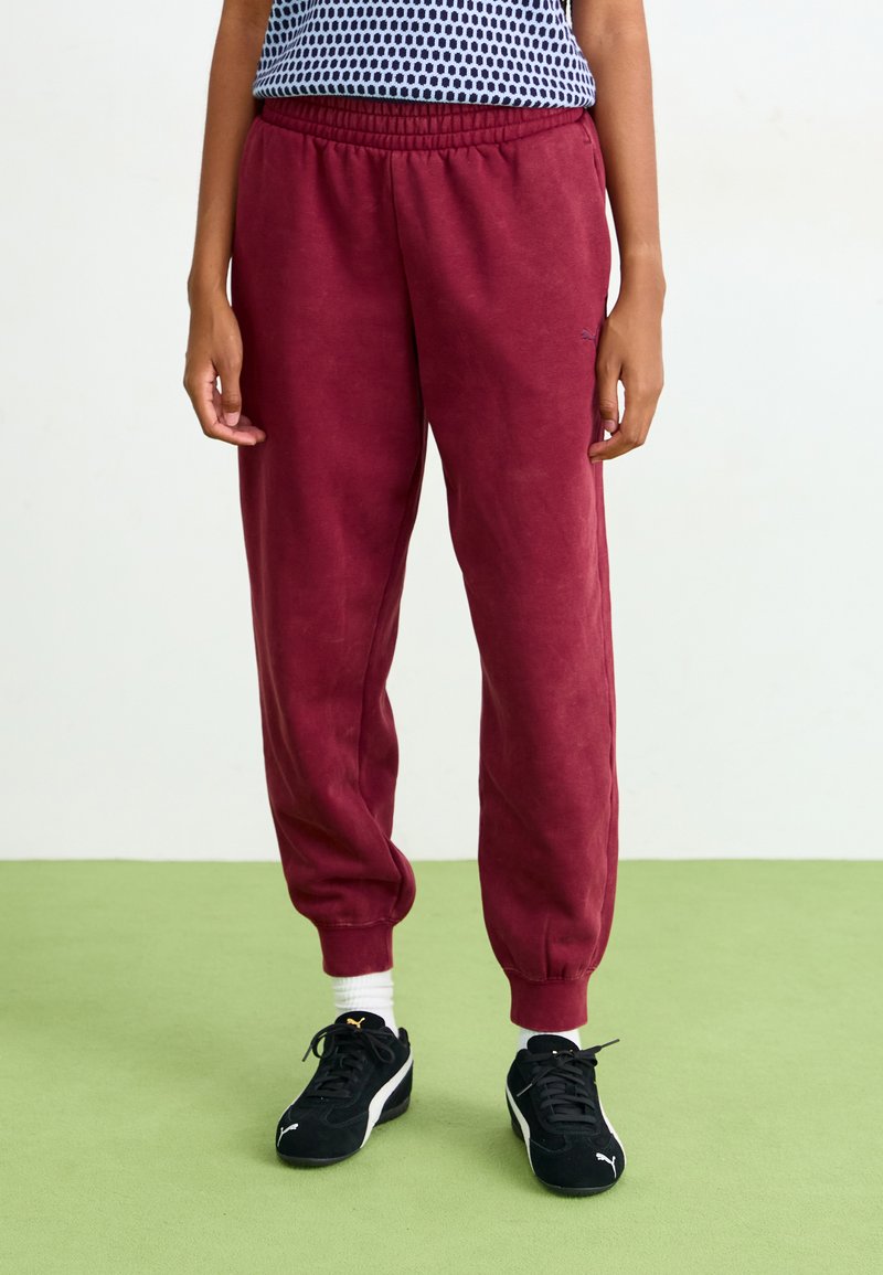 Bordeaux sweatpants med elastisk talje og manchetter, lavet af blødt stof. Sorte træningssko med hvide detaljer, stående på grønne gulve.