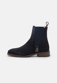 Bota Chelsea de ante azul marino con paneles laterales elásticos, punta redonda y suela de goma marrón. Cuenta con una lengüeta para facilitar su colocación y extracción.