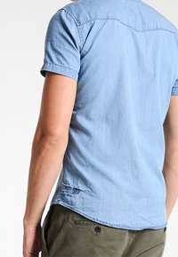 Persona vistiendo una camisa de mezclilla ligera de manga corta y pantalones verdes oliva, vista desde la espalda contra un fondo simple.