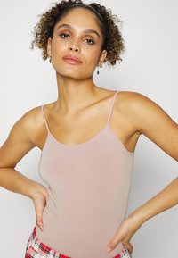 Camiseta de alças finas em rosa claro, feita de um tecido suave e elástico. Modelo posa com uma mão no quadril, vestindo calças de pijama xadrez.