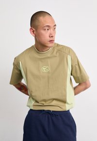 Krátký rukáv tričko v khaki a světle zelené barvě, s kulatým výstřihem a logem Nike Air uprostřed. Hladká bavlněná tkanina.