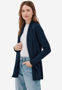 Cardigan bleu marine en tissu doux avec col côtelé, design à ouverture frontale, manches longues, porté sur une chemise blanche et un jean clair.