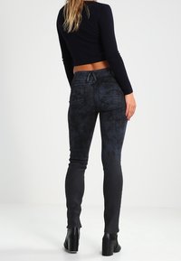 Vaqueros skinnies negros con un patrón desgastado, cintura alta, diseño de cinco bolsillos y detalle en el yugo trasero, combinados con una blusa ajustada de canalé y tacones negros.