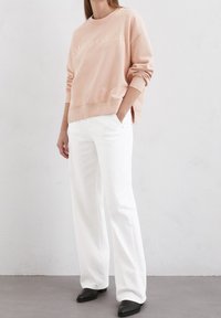 Felpa rosa con logo ricamato, abbinata a pantaloni bianchi a gamba larga e stivaletti neri, su sfondo grigio chiaro.