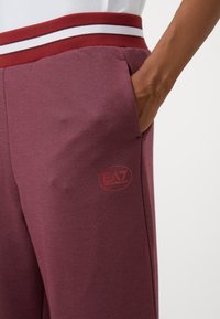 Oseba v maroon hlačah z črtastim rdečim in belim pasom, z roko, ki počiva v sprednjem žepu blizu logotipa EA7.
