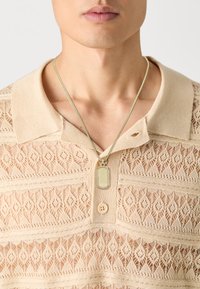 Μπεζ πλεκτό πουκάμισο πόλο με σχέδιο δαντέλας, περίπλοκους διαμαντένιους σχεδιασμούς και μια διακοσμητική κρεμαστή διαθήκη με ετικ έτα.