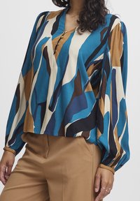 Blusa in teal, marrone e crema con un motivo astratto, dotata di scollo a V e tre bottoni. Maniche lunghe a palloncino con polsini elastici.