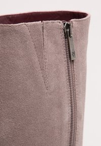 Närbild av en mauve mockaskinshäft med synlig sidodragkedja och vinröd inredning.