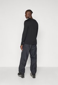 Schwarzes Langarmshirt mit verstellbaren Manschetten, kombiniert mit locker sitzenden blauen Trainingshosen, detaillierten Reißverschlusstaschen und schwarzem Schuhwerk mit Logos.