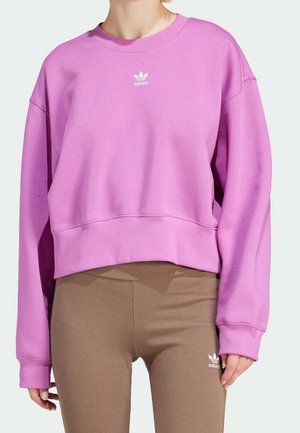 Personne portant un sweat Adidas rose ample et des leggings Adidas marron moulants, tous deux avec des logos Adidas blancs.