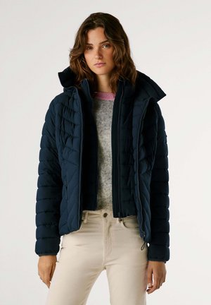 Chaqueta de puffer azul marino con diseño acolchado, cierre de cremallera y capucha. Lleva puesta sobre un suéter gris y combinada con pantalones ajustados de color claro.