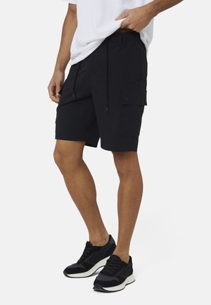 Mężczyzna w czarnych szortach cargo i czarnych trampkach, stojący z jedną ręką poprawiającą białą koszulkę.