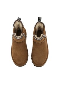 Para brązowych zamszowych kapci typu slip-on z białym futrem wewnętrznym i ciemno-brązowym wykończeniem na białym tle.